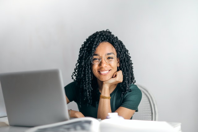 Female black student wearing a dark green shit with glasses smiling while siting at a desk.